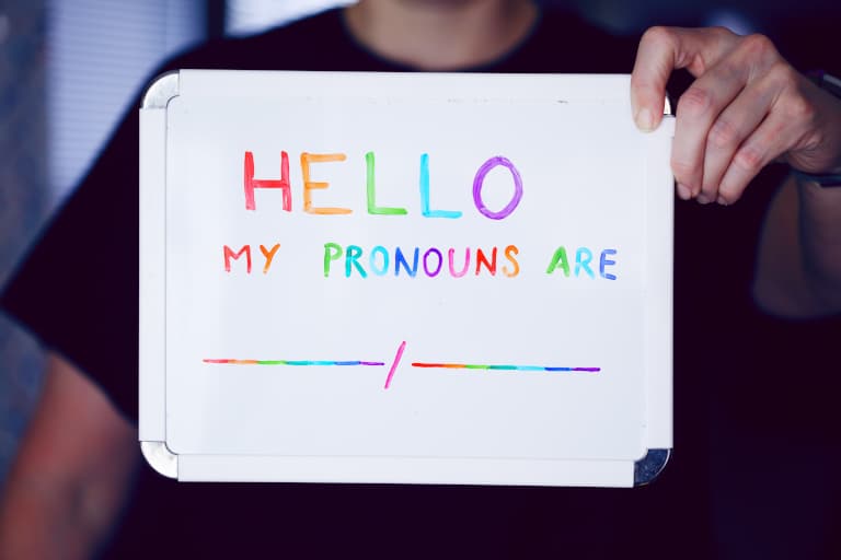 Photo of a person holding a small whiteboard on which it's written in a rainbowy font: “Hello. My pronouns are (blank)/(blank)”